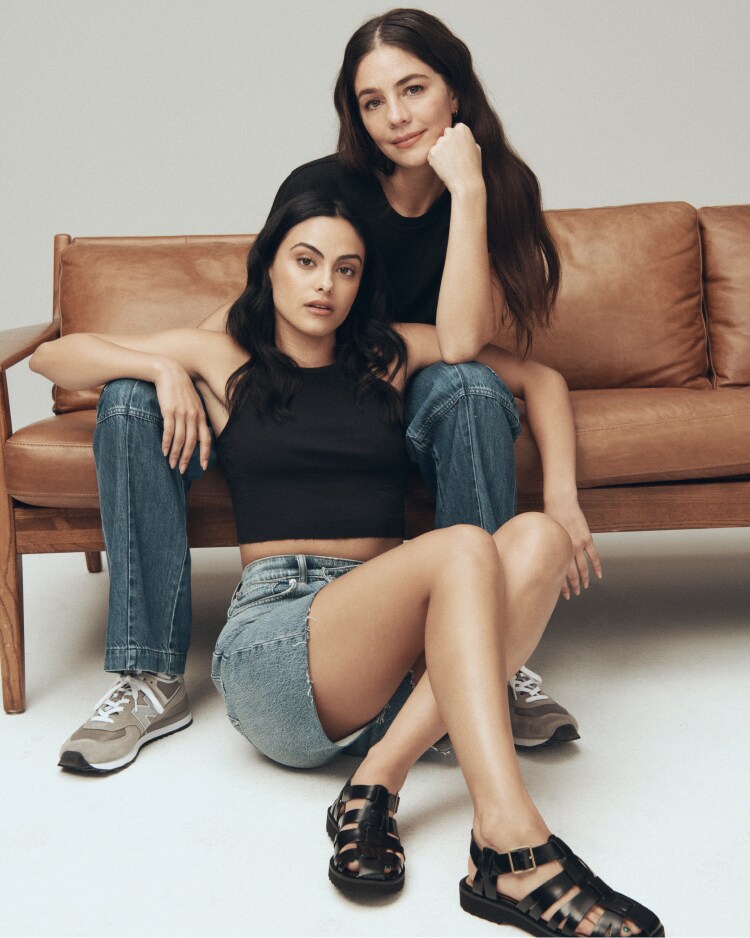 man with long brown hair and headphone sitting on a large speaker in light wash jeans and a blue-grey loose-fitting shirt