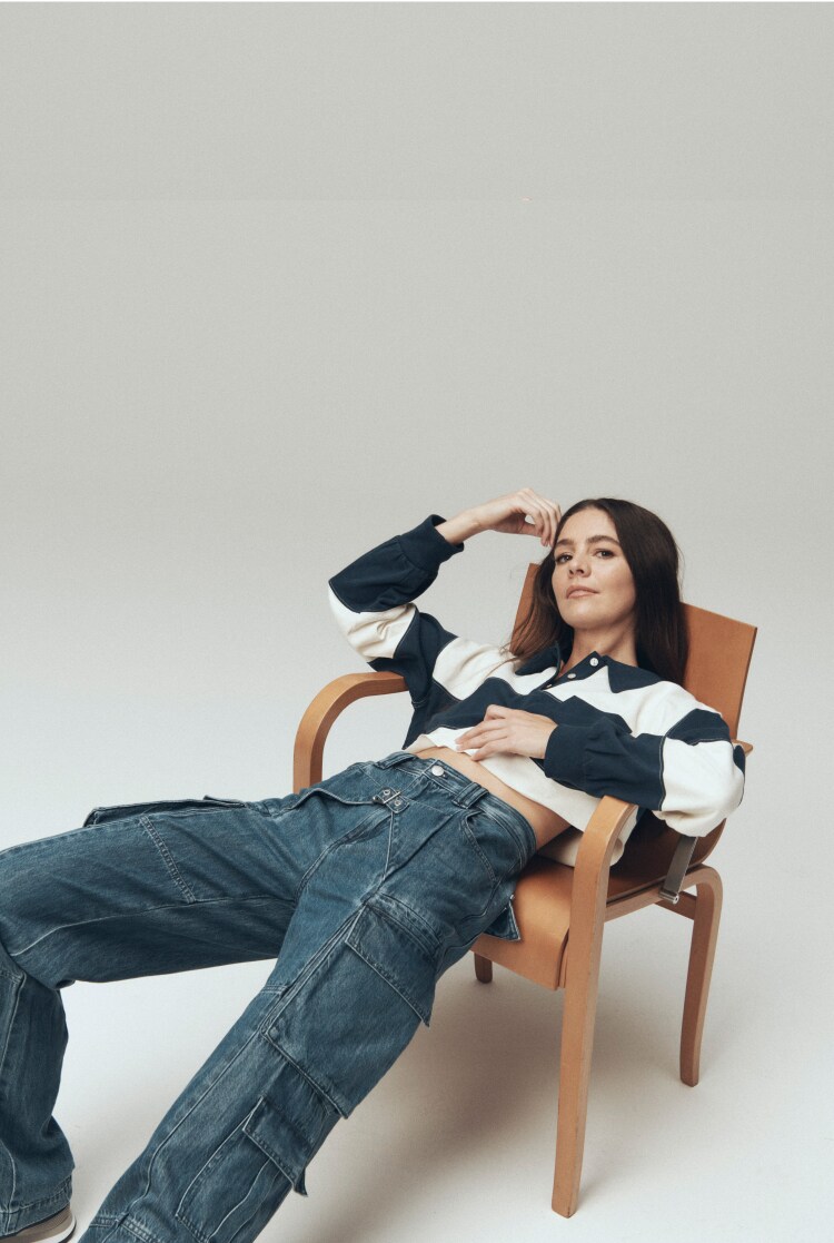 man with long brown hair and headphone sitting on a large speaker in light wash jeans and a blue-grey loose-fitting shirt
